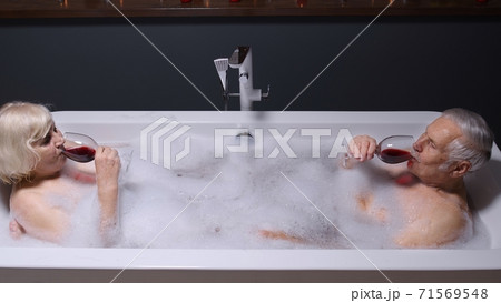 Senior couple man and woman lying in warm bath with bubbles, enjoying relaxation, drinking red wine Senior couple man and woman lying in warm bath with bubbles, enjoying relaxation, drinking red wine 71569548