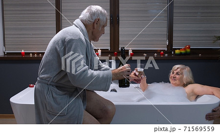 Senior grandmother is taking foamy bath with candles. Grandfather giving glass with champagne 71569559