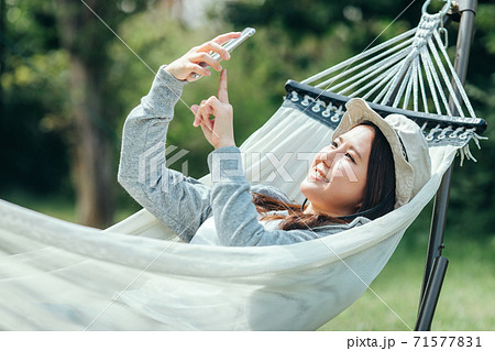 キャンプ場でハンモックに乗りながらスマホで写真を撮るアジア人女性(自撮り) キャンプ場でハンモックに乗りながらスマホで写真を撮るアジア人女性(自撮り) 71577831