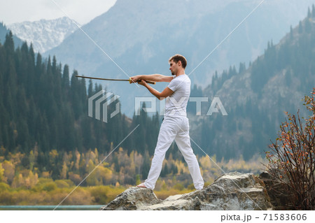A young man stands on a large stone and holds a Japanese sword in his hands. 71583606