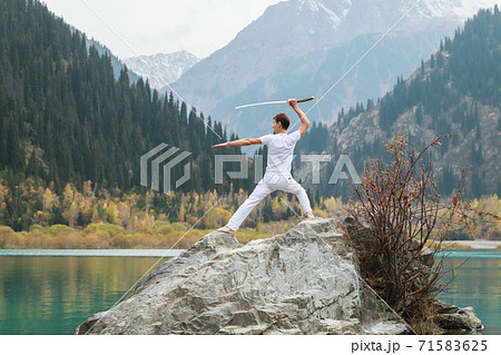 A man in white clothes with a sword stands on a large stone among the highlands. 71583625