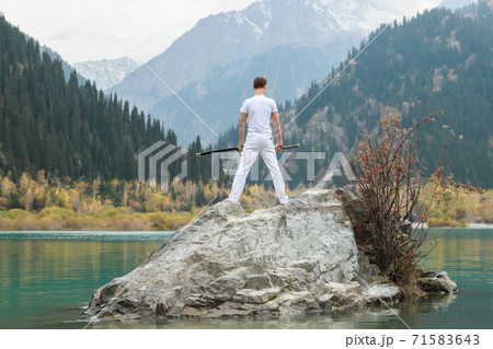 A man in white clothes with a sword stands on a large stone among the highlands. A man in white clothes with a sword stands on a large stone among the highlands. 71583643