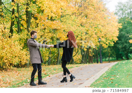 Young red haired woman puts on face mask while walking with young man in autumn park. Young red haired woman puts on face mask while walking with young man in autumn park. 71584997
