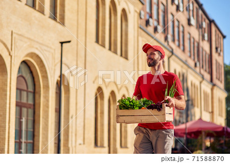 Young cheerful caucasian male courier in uniform looking aside and smiling while delivering wooden box with fresh green grocery, standing on sunny street 71588870