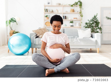 Sports and pregnancy concept. Lovely black expectant woman hugging her big belly while meditating on mat at home 71601663