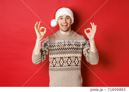 Portrait of happy and pleased handsome man, wearing winter sweater and santa hat, showing okay signs, praise good christmas party, standing over red background Portrait of happy and pleased handsome man, wearing winter sweater and santa hat, showing okay signs, praise good christmas party, standing over red background 71609865