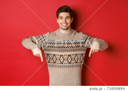 Concept of christmas celebration, winter holidays and lifestyle. Attractive happy man in xmas sweater showing promotion, pointing fingers down at logo, standing over red background 71609884