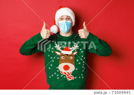 Concept of new year, covid-19 and social distancing. Cheerful handsome man in santa hat and medical mask, showing thumbs-up, enjoying christmas party, standing over red background Concept of new year, covid-19 and social distancing. Cheerful handsome man in santa hat and medical mask, showing thumbs-up, enjoying christmas party, standing over red background 71609945