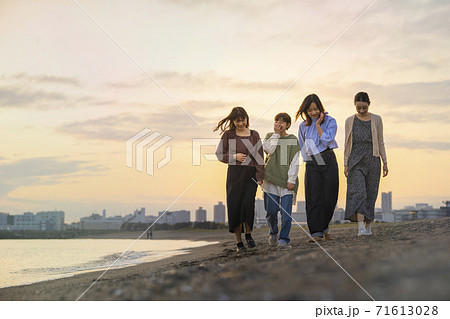 夕暮れの中、寄り添って海沿いを歩く女性4人 71613028