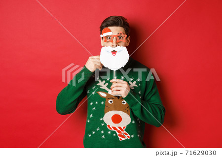 Portrait of surprised man in green christmas sweater, holding funny santa claus mask, celebrating new year, standing over red background 71629030