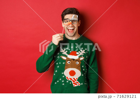 Portrait of amused handsome man in christmas sweater, holding funny party mask and smiling, celebrating winter holidays, standing over red background Portrait of amused handsome man in christmas sweater, holding funny party mask and smiling, celebrating winter holidays, standing over red background 71629121
