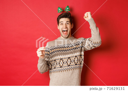 Cheerful guy with a sparkler rejoicing and wishing happy new year, raising hand up and shouting for joy, standing over red background Cheerful guy with a sparkler rejoicing and wishing happy new year, raising hand up and shouting for joy, standing over red background 71629131