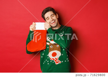 Portrait of happy attractive man in green sweater, looking delighted to receive gifts in christmas stocking, standing over red background 71629800