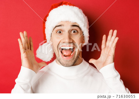 Close-up of happy young man in santa hat, making big christmas announcement, smiling amazed, standing over red background 71630055