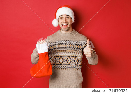 Concept of winter holidays, new year and celebration. Cheerful handsome man in santa hat and sweater, showing christmas stocking with candies and gifts, making thumbs-up 71630525