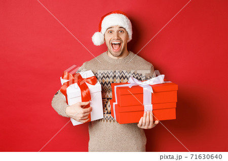 Concept of winter holidays, new year and celebration. Image of amazed and happy man in sweater and santa hat, holding christmas gifts and shouting for joy, red background Concept of winter holidays, new year and celebration. Image of amazed and happy man in sweater and santa hat, holding christmas gifts and shouting for joy, red background 71630640