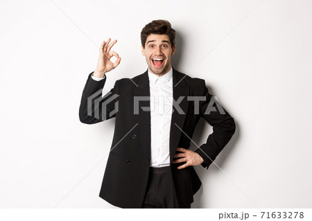 Concept of new year party, celebration and lifestyle. Portrait of pleased handsome guy in black suit, praise something good, showing okay sign in approval, standing over white background Concept of new year party, celebration and lifestyle. Portrait of pleased handsome guy in black suit, praise something good, showing okay sign in approval, standing over white background 71633278