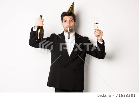 Concept of holidays and lifestyle. Handsome guy celebrating birthday, blowing party whistle and holding champagne, saying a toast, standing in suit over white background 71633281