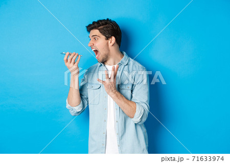Angry and annoyed guy screaming at mobile phone, shouting on speakerphone while standing over blue background Angry and annoyed guy screaming at mobile phone, shouting on speakerphone while standing over blue background 71633974