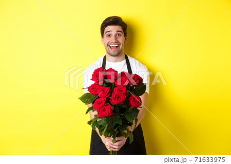 Seller in flower shop wearing black apron, giving bouquet of roses and smiling, standing over yellow background 71633975