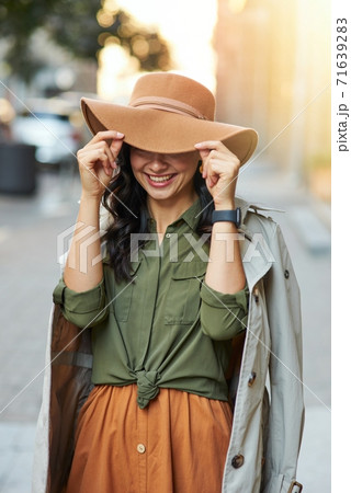 Playful beauty. Young happy caucasian woman adjusting hat and smiling while standing on the city street, enjoying walking on warm autumn day Playful beauty. Young happy caucasian woman adjusting hat and smiling while standing on the city street, enjoying walking on warm autumn day 71639283