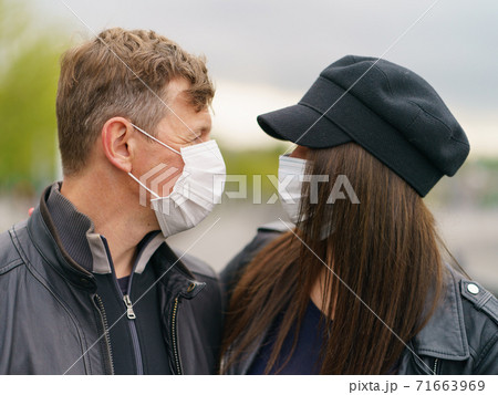Hugging heterosexual couple in love of young handsome man and pretty woman in rainy spring day. They look at each other. Wearing of protective face masks during coronavirus pandemic. Hugging heterosexual couple in love of young handsome man and pretty woman in rainy spring day. They look at each other. Wearing of protective face masks during coronavirus pandemic. 71663969