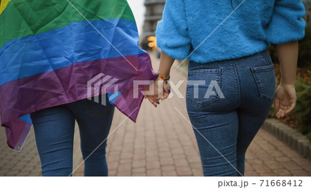 Backside view: Female lesbian couple holds hands wearing rainbow flag 71668412