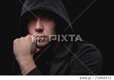 Portrait of a large attractive man in a black hood holds his chin with his hand in a pensive pose. Secretive face in the shadow of the outlaw 71668413
