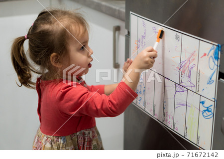 Childhood, infants concept - Little blonde minor preschool girl 2-3 years old in dress draws with marker on planning board on refrigerator in kitchen. Baby toddler get creative on fridge with magnets Childhood, infants concept - Little blonde minor preschool girl 2-3 years old in dress draws with marker on planning board on refrigerator in kitchen. Baby toddler get creative on fridge with magnets 71672142