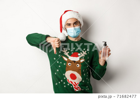 Concept of covid-19 and Christmas holidays. Disappointed man in face mask and santa hat, dislike hand sanitizer, showing thumbs down, standing over white background 71682477