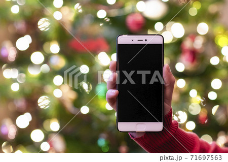 Woman holding mobile phone and showing screen with christmas tree lights on the background. Mockup for greeting. Concept. 71697563