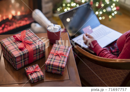 Woman holding credit card using laptop for making order sitting at table with packaging gift, cup of cocoa and marshmallows near fireplace and christmas tree. Online shopping concept Woman holding credit card using laptop for making order sitting at table with packaging gift, cup of cocoa and marshmallows near fireplace and christmas tree. Online shopping concept 71697757