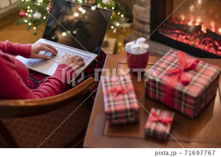Christmas. Woman using laptop for searching gift ideas sitting at table with ready gift boxes and cup of cocoa and marshmallows near fireplace and christmas tree. Concept 71697767