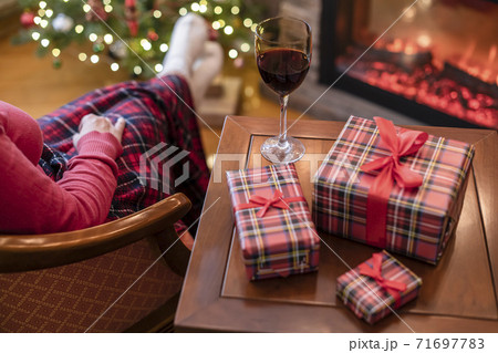 Alone woman covered plaid with glass of wine sitting and relaxation on armchair near fareplace and christmas tree after finishing pakking gift boxes for family. 71697783