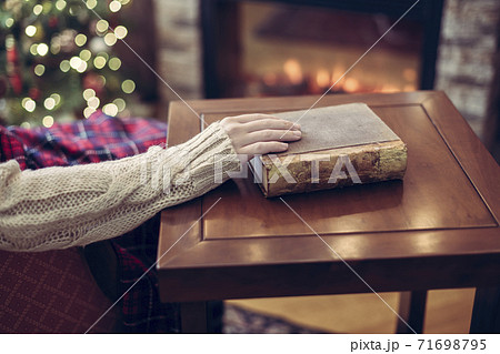 Woman hand with old retro book on wooden table near christmas tree and fireplace. Toned Woman hand with old retro book on wooden table near christmas tree and fireplace. Toned 71698795