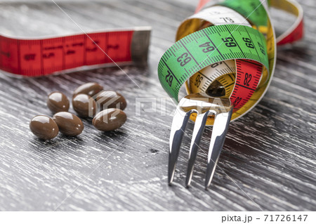 A shiny fork wrapped in a multi-colored centimeter to measure body shapes and brown medical capsules on a black old wooden table. The concept of nutritional supplements for weight loss A shiny fork wrapped in a multi-colored centimeter to measure body shapes and brown medical capsules on a black old wooden table. The concept of nutritional supplements for weight loss 71726147