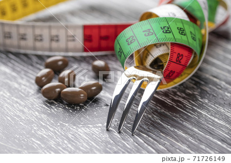 A shiny fork wrapped in a multi-colored centimeter to measure body shapes and brown medical capsules on a black old wooden table. The concept of nutritional supplements for weight loss A shiny fork wrapped in a multi-colored centimeter to measure body shapes and brown medical capsules on a black old wooden table. The concept of nutritional supplements for weight loss 71726149