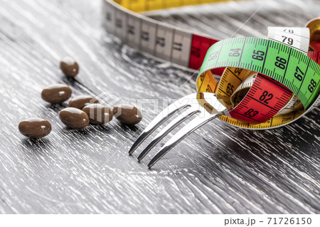 A shiny fork wrapped in a multi-colored centimeter to measure body shapes and brown medical capsules on a black old wooden table. The concept of nutritional supplements for weight loss A shiny fork wrapped in a multi-colored centimeter to measure body shapes and brown medical capsules on a black old wooden table. The concept of nutritional supplements for weight loss 71726150