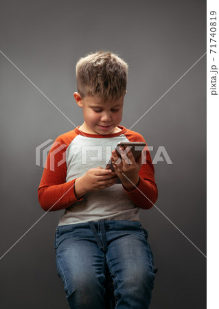 little boy in red shirt looking at the smartphone holding it in his hands and smile isolated on grey background. Human emotions, facial expression concept. Facial expressions, emotions, feelings little boy in red shirt looking at the smartphone holding it in his hands and smile isolated on grey background. Human emotions, facial expression concept. Facial expressions, emotions, feelings 71740819