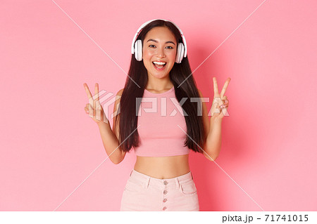 Portrait of beautiful and upbeat, smiling asian girl listening music in headphones, showing peace kawaii gesture, sending positive vibes, standing over pink background 71741015