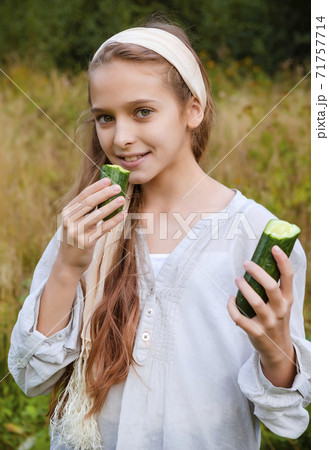 1 white girl 11 years old in a white shirt eating a fresh cucumber on the background of grass 71757714