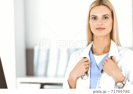 Blond female physician is standing at her workplace near desktop computer. Woman-doctor is excited and happy of her profession. Medicine concept 71760780