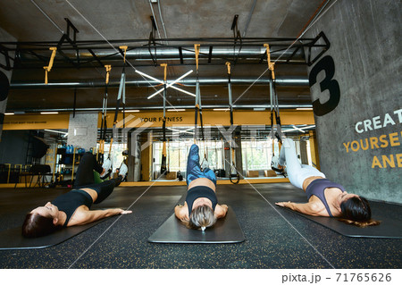 Three young caucasian athletic women in sportswear lying on the floor and exercising with fitness trx straps at industrial or crossfit gym 71765626