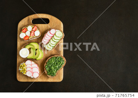 Various homemade sandwiches with vegetables radishes, cucumbers, avocado on slices of dark bread on cutting board. Vegetarian food flat lay on dark concrete background with place for text. Various homemade sandwiches with vegetables radishes, cucumbers, avocado on slices of dark bread on cutting board. Vegetarian food flat lay on dark concrete background with place for text. 71765754