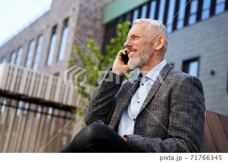 Portrait of elegant mature businessman talking on the phone while taking a break, sitting on the bench outdoors Portrait of elegant mature businessman talking on the phone while taking a break, sitting on the bench outdoors 71766345