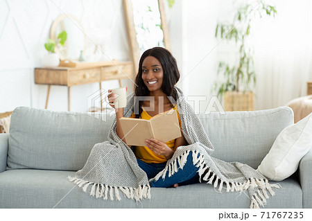 Lovely black woman reading book, covered with warm plaid, drinking hot aromatic coffee on sofa at home Lovely black woman reading book, covered with warm plaid, drinking hot aromatic coffee on sofa at home 71767235