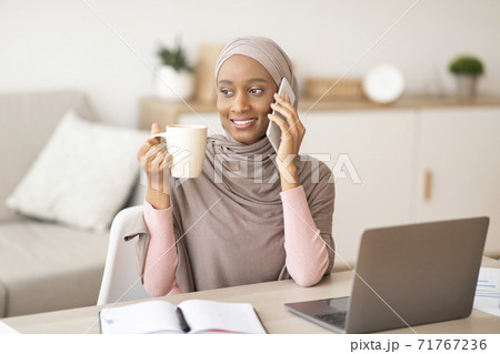 Lovely black lady in hijab talking on smartphone and drinking coffee near laptop computer at home Lovely black lady in hijab talking on smartphone and drinking coffee near laptop computer at home 71767236