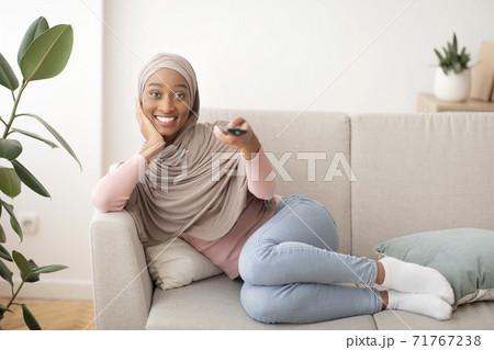 Positive black woman in hijab watching TV on couch at home, copy space Positive black woman in hijab watching TV on couch at home, copy space 71767238