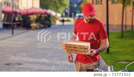 Young caucasian delivery man in red uniform delivering pizza, standing near his scooter 71771322