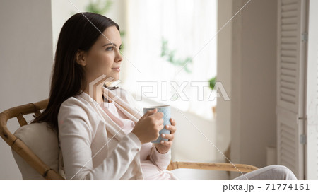 Tranquil woman sitting in armchair by window drinking tea relaxing Tranquil woman sitting in armchair by window drinking tea relaxing 71774161